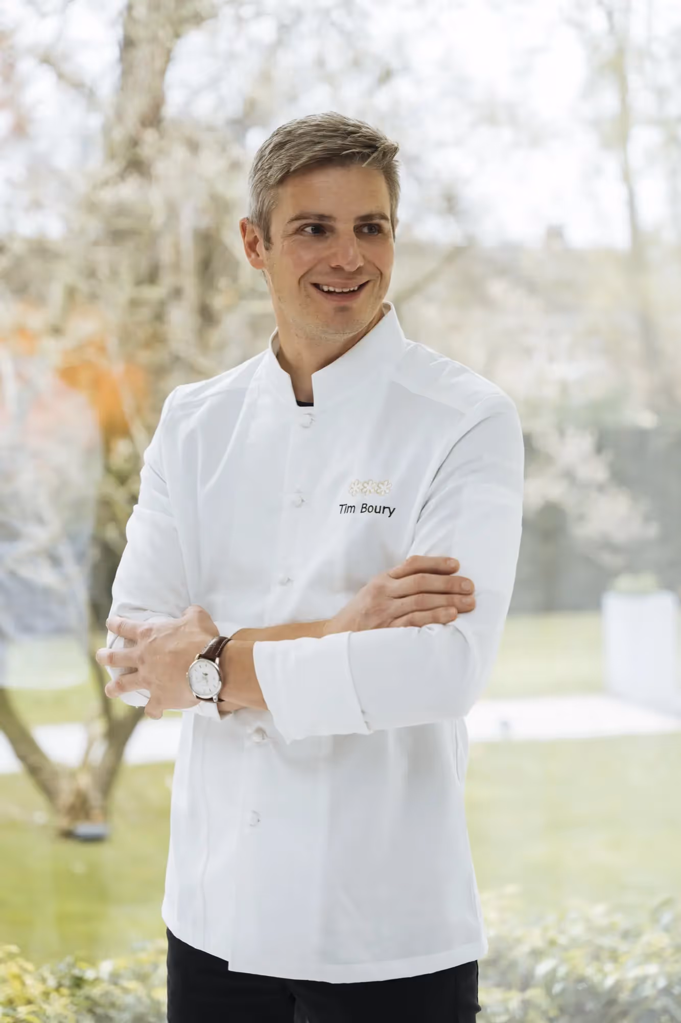 Tim boury voor een raam met zicht op een groene tuin, gekleed in een witte koksvest met gekruiste armen.