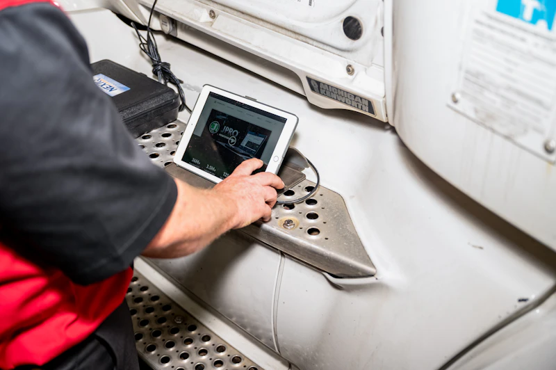 Diagnostics technician using a tablet to perform electronic testing on a semi-truck. The tablet screen displays "IPRO."
