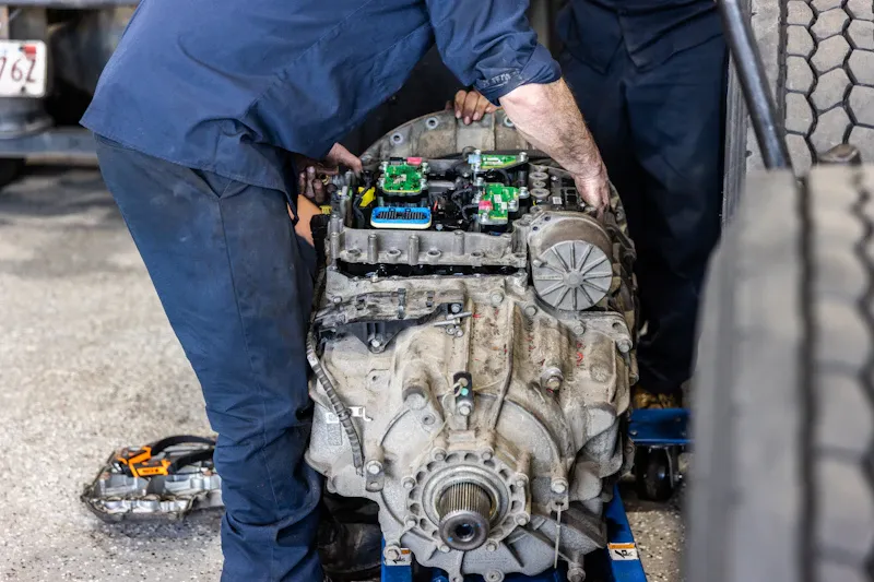 Heavy-duty transmission repair with two technicians handling a removed gearbox and exposed electronic control modules.