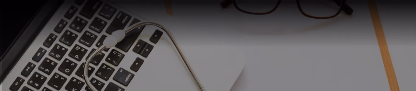 Stethoscope resting on a laptop keyboard with a pair of glasses nearby on a desk.