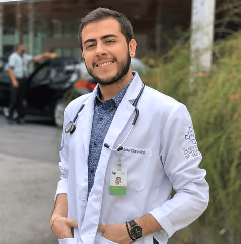 Smiling male doctor wearing a white coat with stethoscope and ID badge, standing outdoors.