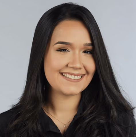 Smiling woman with long dark hair wearing a black top and a pendant necklace against a light gray background.