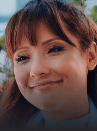 Close-up of a smiling woman with reddish-brown hair and bangs outdoors.
