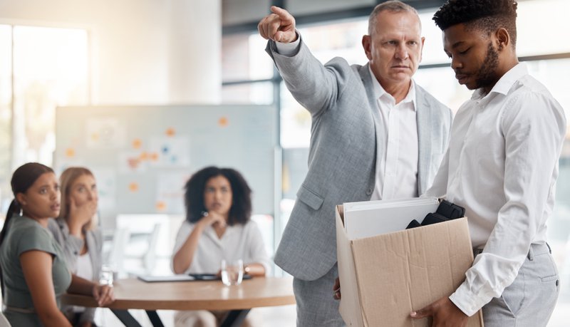 Black man, sad and fired by boss in a meeting holding a box in disappointment at the office. Manager or company leader pointing to the exit and firing employee in front of colleagues at the workplace, Discrimination