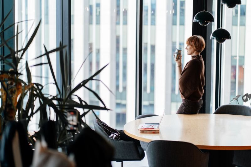 Employee standing alone at office window holding coffee, reflecting on a hostile work environment at work