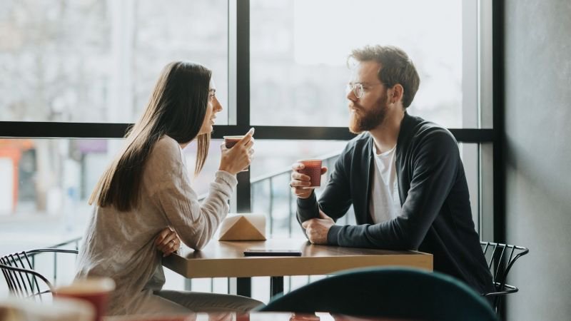 Two people having a candid conversation over coffee at a café table near a window