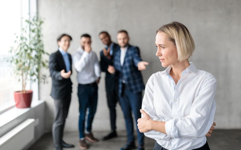 A woman stands apart from a group of male coworkers in an office, who appear to be talking about her behind her back