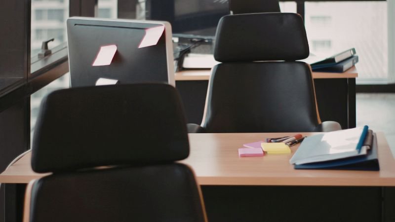An empty office desk with chairs and a laptop, lit by warm natural light from a window
