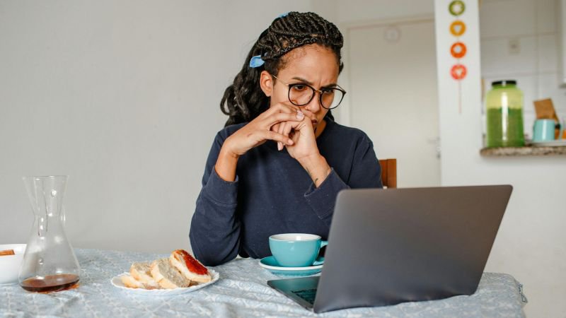 A woman with glasses sits at her kitchen table, leaning forward with a focused and worried expression while researching the statute of limitations for her California employment claim on a laptop.