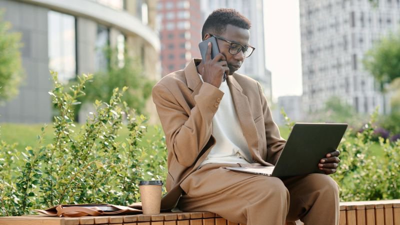 A man in a tan blazer sits outdoors on a bench, talking on the phone while reviewing his laptop, representing an employee taking action after learning about the statute of limitations on his California employment claim.