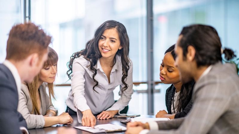 Diverse group of five professionals gathered around a conference table, woman in center leaning forward and engaging collaboratively with colleagues in a bright modern office