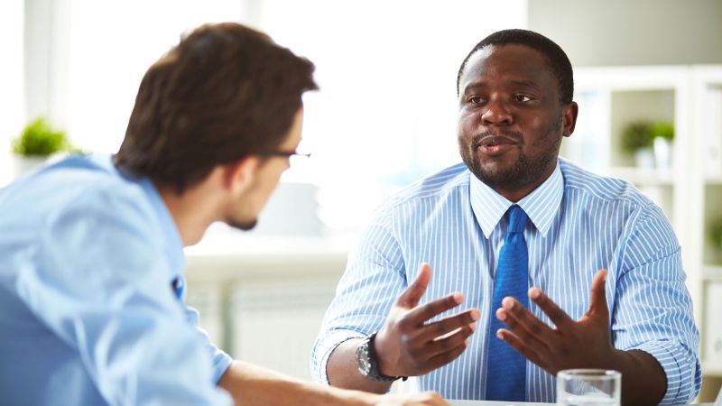 Two professionals in a bright office having a focused conversation, one man gesturing expressively while explaining employment rights to the other