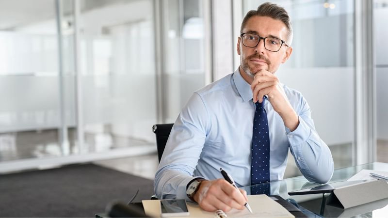 Professional man in dress shirt and tie sitting at a glass desk, looking thoughtfully away from camera while holding a pen