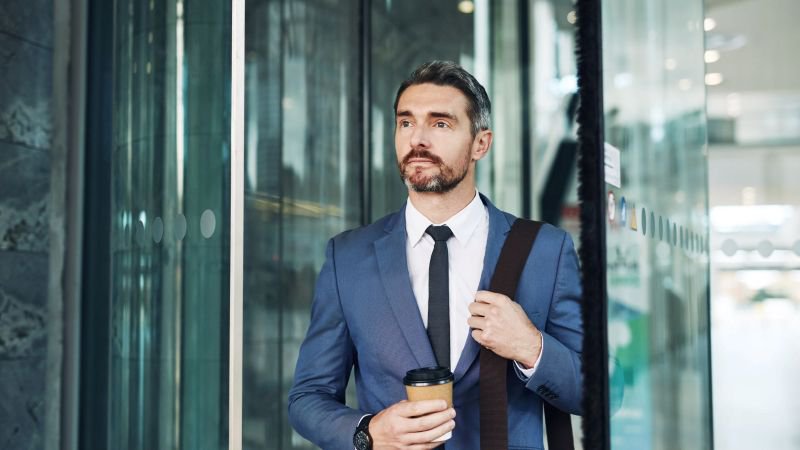 Professional man in blue suit holding a coffee cup and bag, standing outside modern glass office building doors with a composed, forward-looking expression