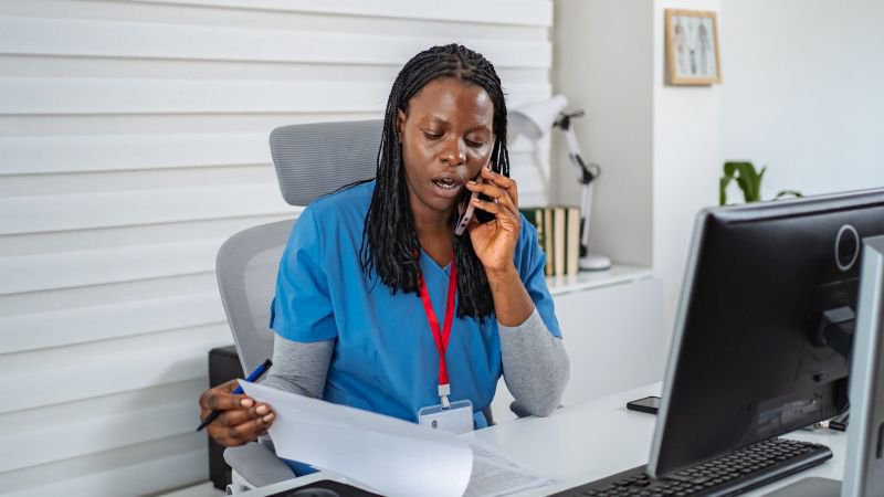 Home health employee in scrubs reviewing documents while on a phone call, representing misclassified caregivers in California