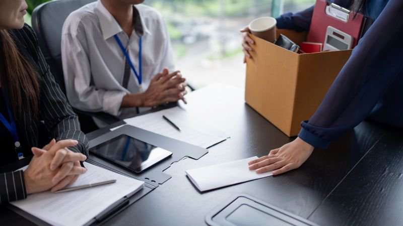 Employee facing forced resignation at a meeting, holding a box of personal belongings