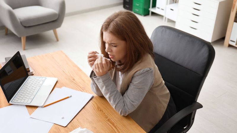 Female employee coughing into a tissue at her office desk, illustrating the onset of a serious health condition that may qualify for FMLA or CFRA protected leave in California
