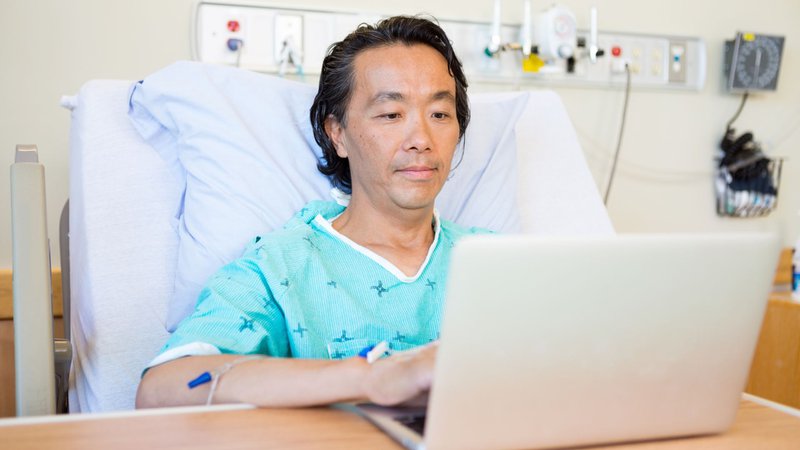 Male patient in a hospital bed using a laptop while receiving IV treatment, representing a California employee on FMLA or CFRA protected leave facing employer pressure to return
