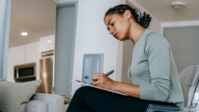 A woman seated at home writing notes on a clipboard, representing a California employee documenting incidents of workplace sexual harassment to support a legal claim