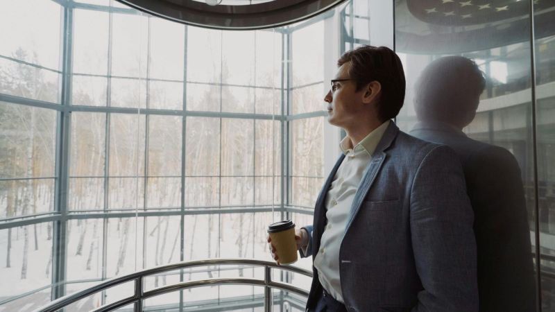 Employee standing at floor-to-ceiling office windows, holding coffee, looking out contemplatively