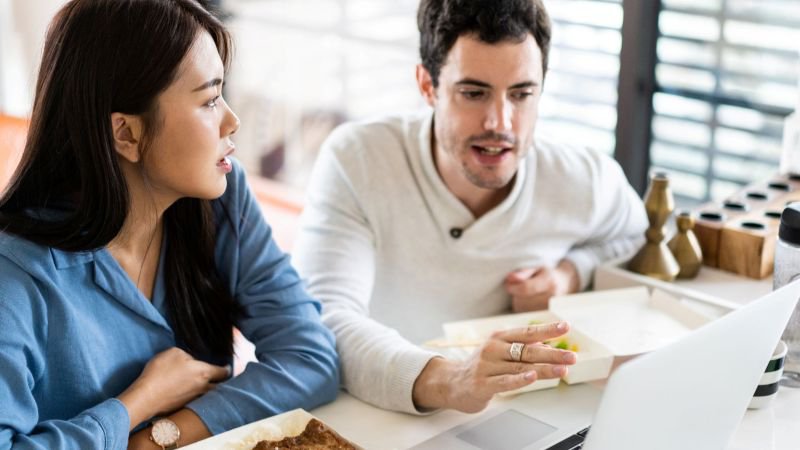 Two employees eating lunch while reviewing work on a laptop, a scenario that triggers meal break premium pay under California law