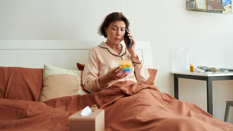 Woman in pajamas sitting in bed on the phone while managing medication, representing a sick day at home under California sick leave law