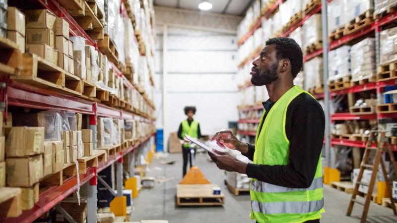 Warehouse employee in a yellow safety vest reviewing inventory on a shelf inside an industrial facility, with a second employee working in the background