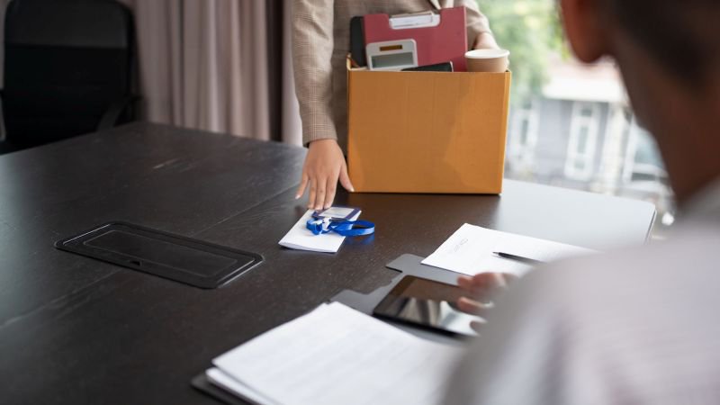 Employee placing ID badge on desk while holding a cardboard box of belongings after being let go from a job in California