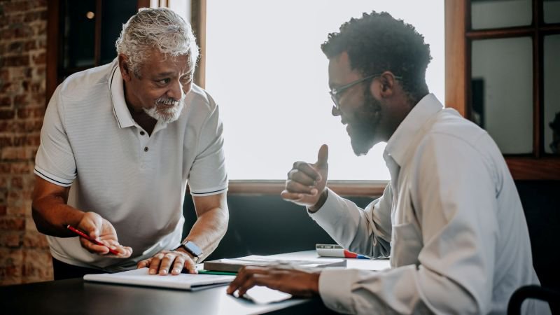 Older professional reviewing documents in discussion with a younger colleague at a workplace table