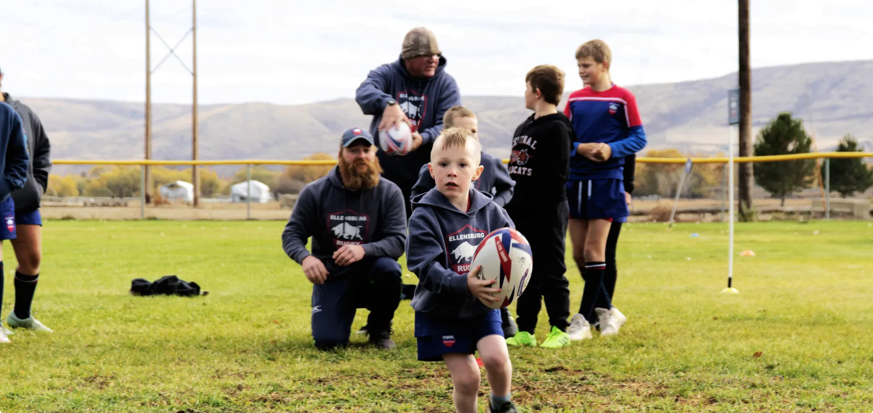 Young boy running with a rugby ball on a grassy field with coaches and other children in the background.