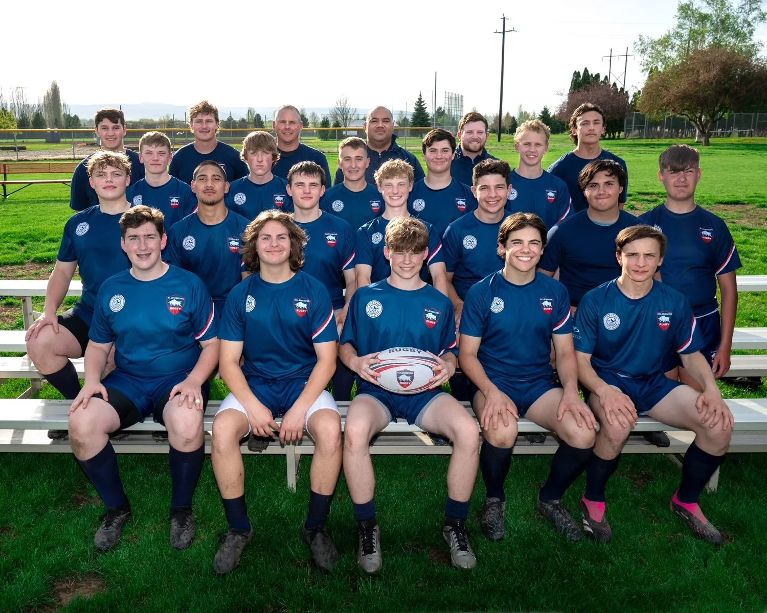 High school boys rugby team in blue uniforms posing on bleachers outdoors with one player holding a rugby ball.