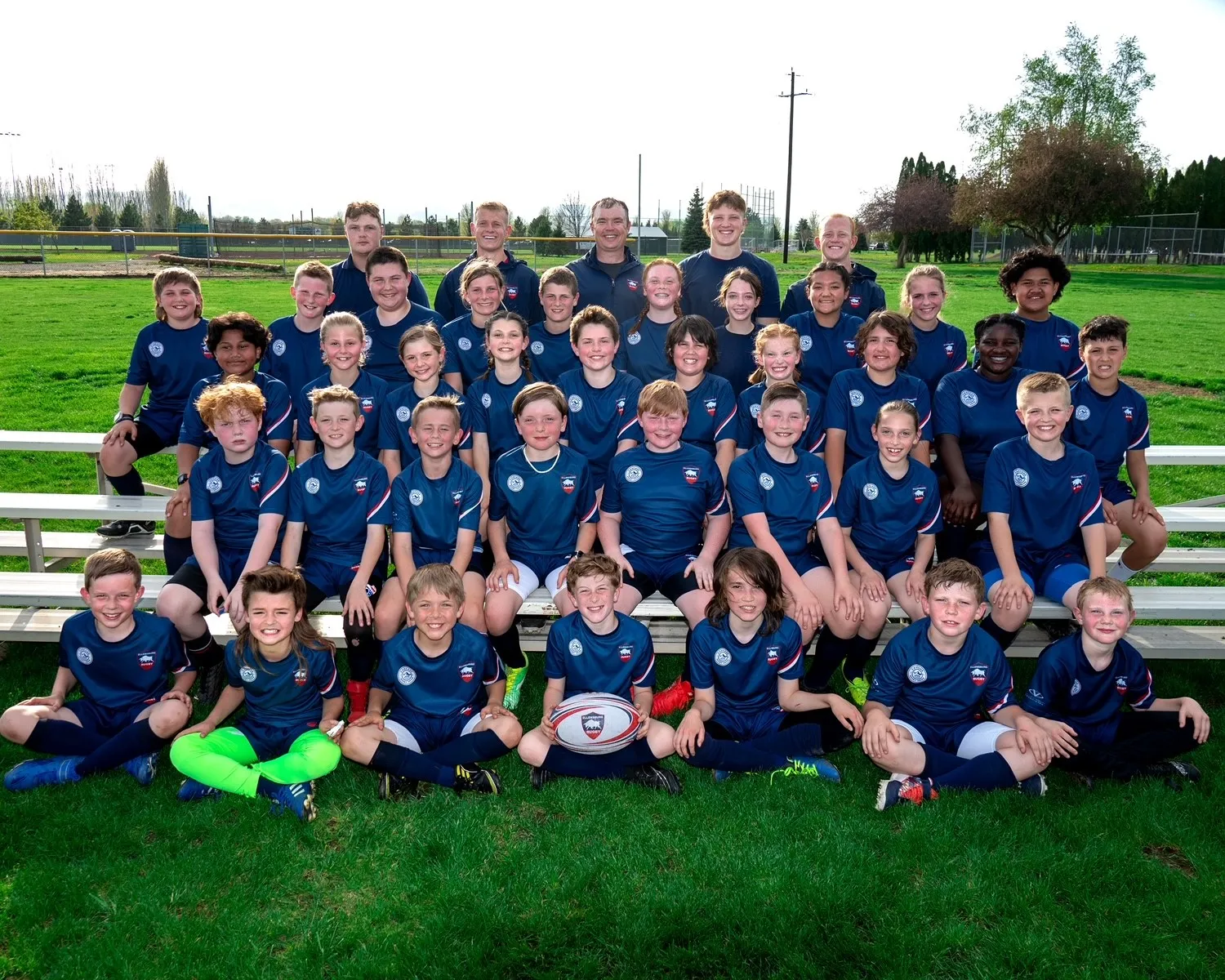 Youth rugby team wearing navy blue uniforms posing outdoors on grass in front of bleachers with a rugby ball.