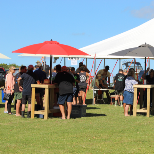 People gathered outdoors around tables under red and gray umbrellas with a white tent in the background on a sunny day.