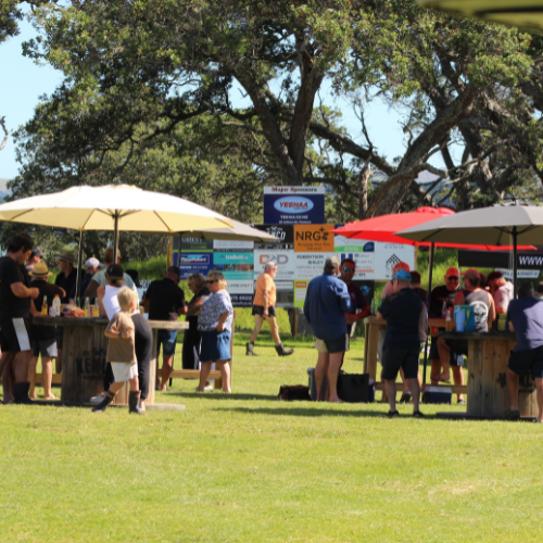 Group of people socialising outdoors under large umbrellas on a sunny day with trees and sponsor signs in the background.