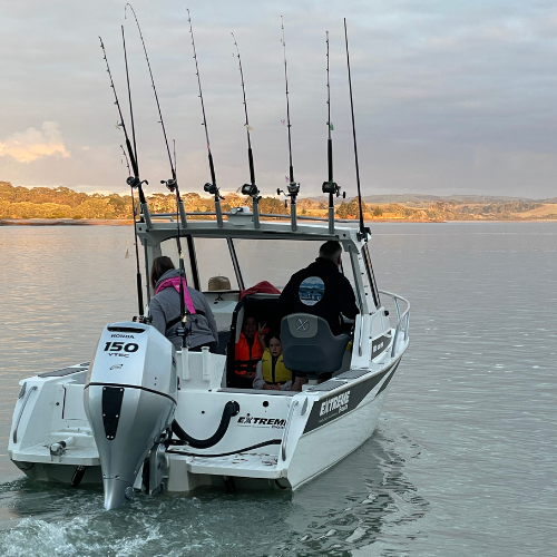 White fishing boat with five fishing rods on the back, two adults and two children onboard on calm water during daytime.