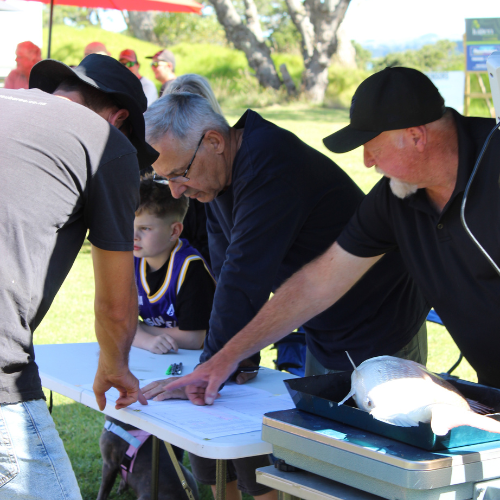 Three men and a boy gathered around a table outdoors, with one man pointing to documents and a fish on a scale nearby.