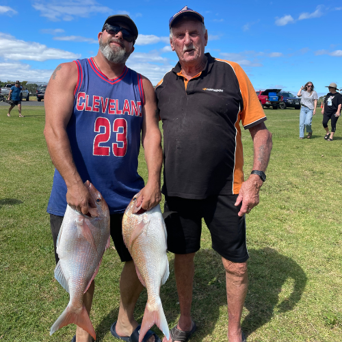 Two men standing on grass holding large fish, with cars and people visible in the background under a blue sky.