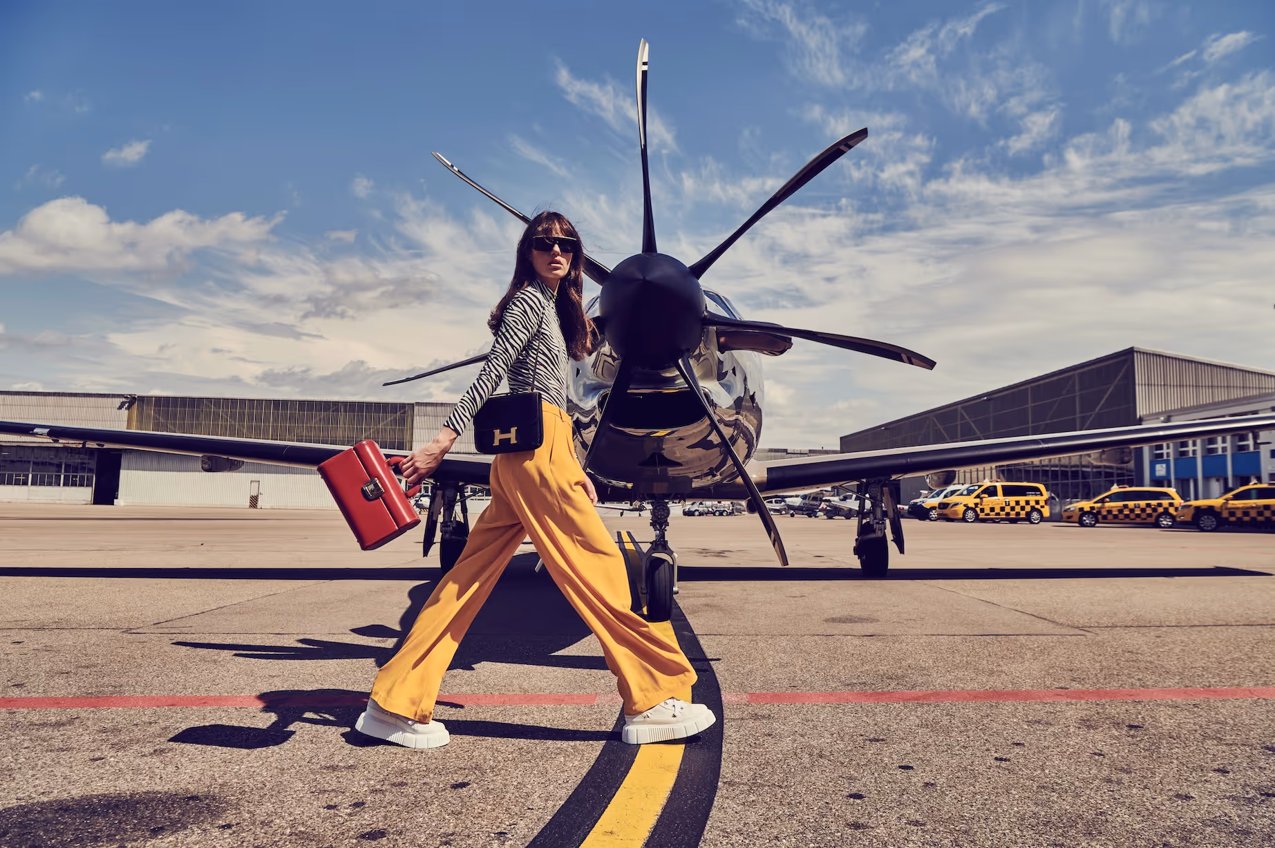 Fashionable woman walking on airport tarmac in front of a Pilatus aircraft, holding red suitcase.