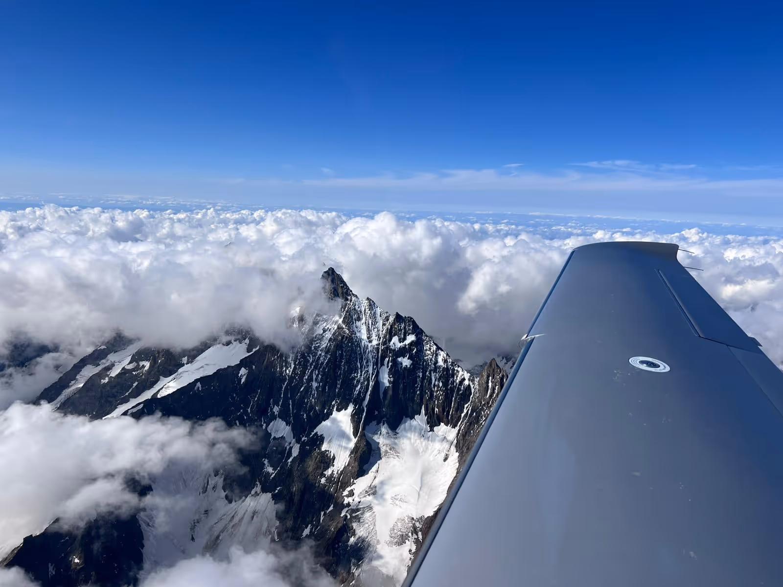 Snow-covered mountain peak seen from airplane wing above the clouds.
