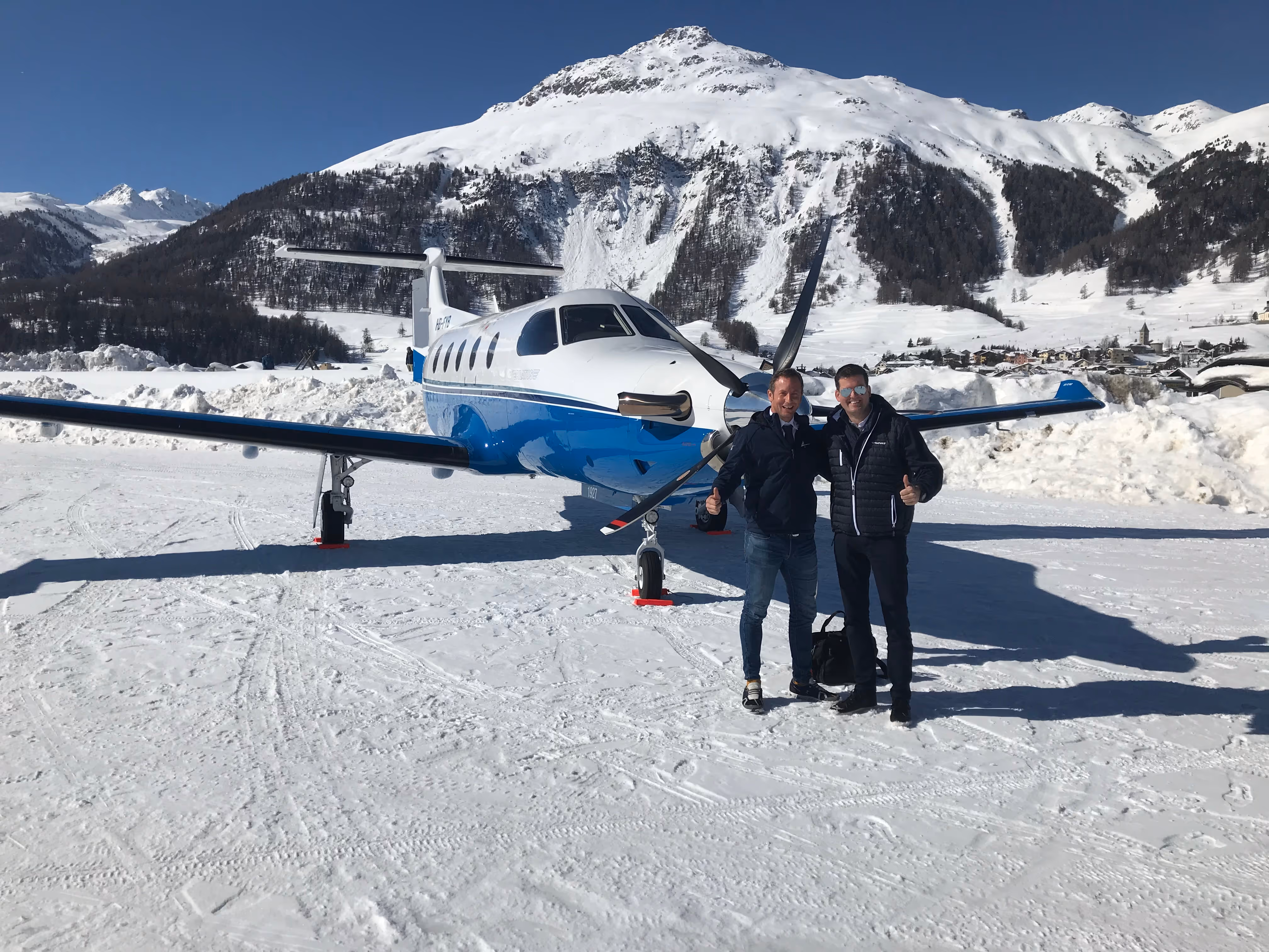 Two pilots standing in front of a Pilatus PC-12 aircraft on snowy runway in the Swiss Alps.