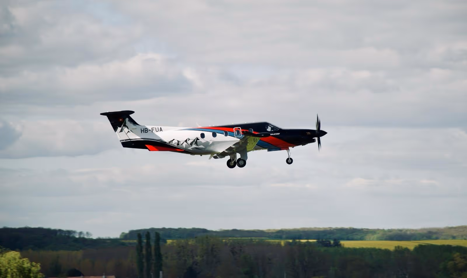 Pilatus PC-12 aircraft with custom livery flying low on approach over green landscape.