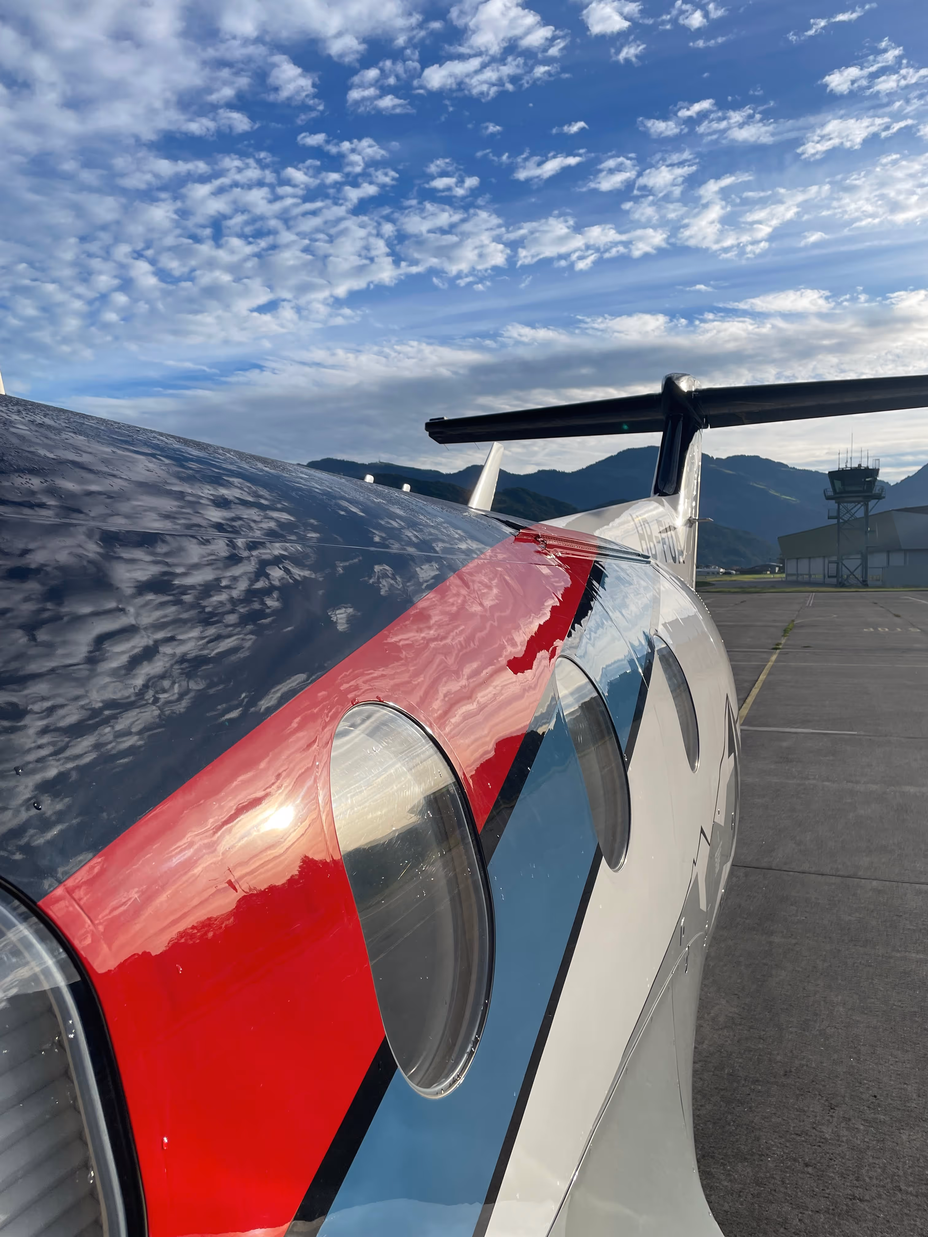 Close-up of Pilatus aircraft fuselage with red, black, and blue livery under cloudy sky.