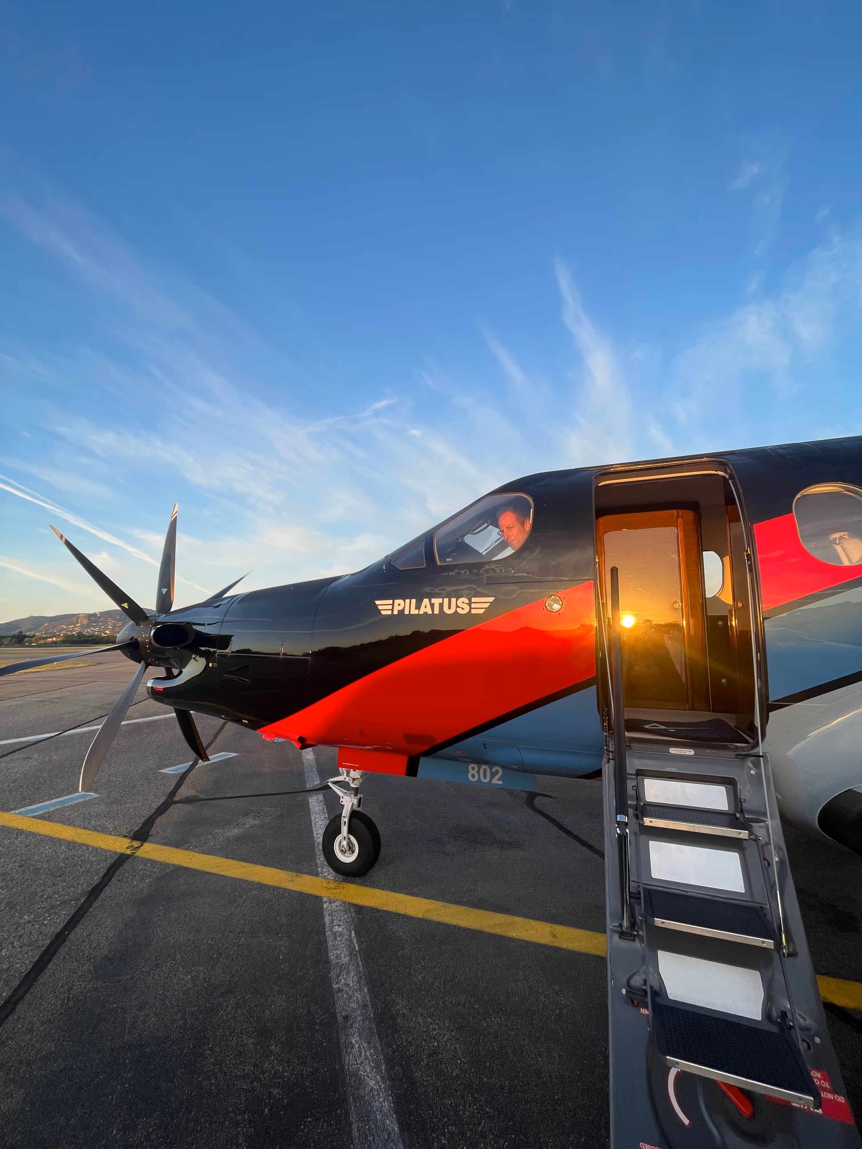 Pilatus aircraft with open door and pilot in cockpit at sunrise on airport tarmac.
