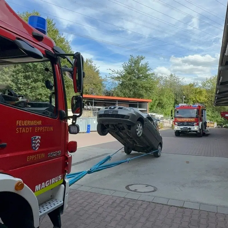 Zugmaschine der Feuerwehr zieht ein Auto, das auf der Rückseite über eine Zugvorrichtung angehoben wird, vor einer Lagerhalle mit einem weiteren Feuerwehrfahrzeug im Hintergrund.
