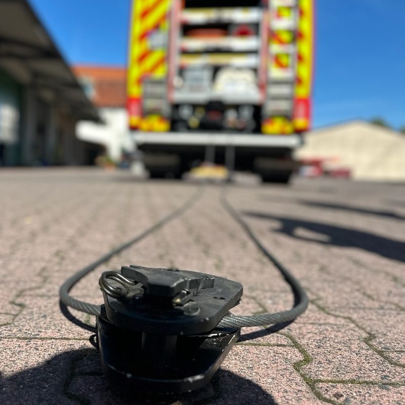 Nahaufnahme einer maschinellen Zugeinrichtung auf Pflastersteinen mit einem Feuerwehrfahrzeug im Hintergrund.