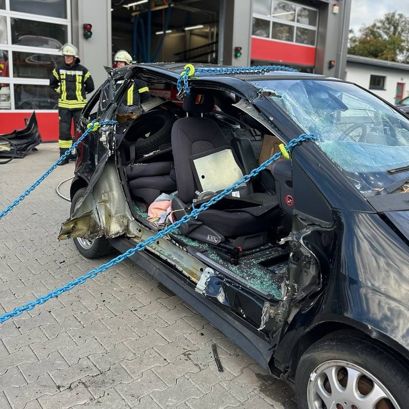 Beschädigtes schwarzes Auto mit zerbrochenem Fenster und blauen Ketten zur Stabilisierung, Feuerwehrleute im Hintergrund vor Feuerwehrgarage. Kettenrettung – Oslo-Methode