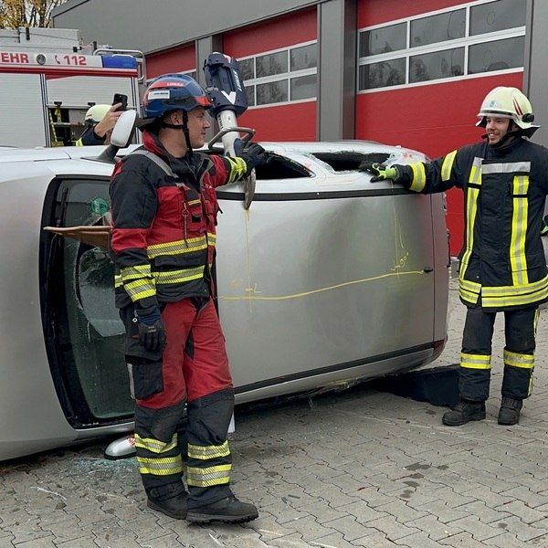 Zwei Feuerwehrleute in Schutzkleidung neben auf der Seite liegendem Auto bei einer Übung zur Unfallrettung. PKW Unfallrettung Seminar