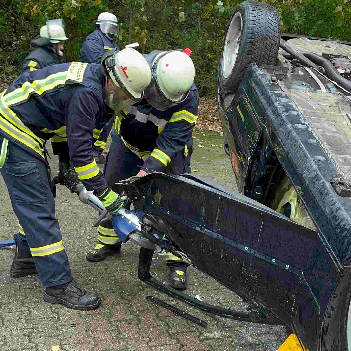 Feuerwehrleute in Schutzkleidung führen eine Unfallrettungsübung an einem auf der Seite liegenden Auto durch. PKW Unfallrettung Seminar