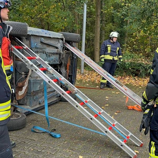 Feuerwehrleute stabilisieren ein auf die Seite gekipptes Auto mit Leitern und Gurten im Außenbereich mit herbstlichem Laub. PKW Unfallrettung Seminar