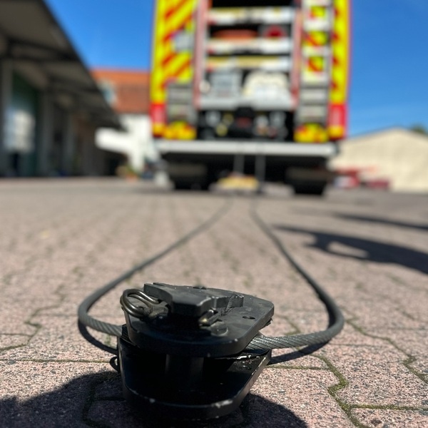 Nahaufnahme einer schwarzen Umlenkrolle mit Stahlseil auf einem gepflasterten Boden vor einem geparkten Feuerwehrfahrzeug. Maschinelle Zugeinrichtung Trainingsseminar
